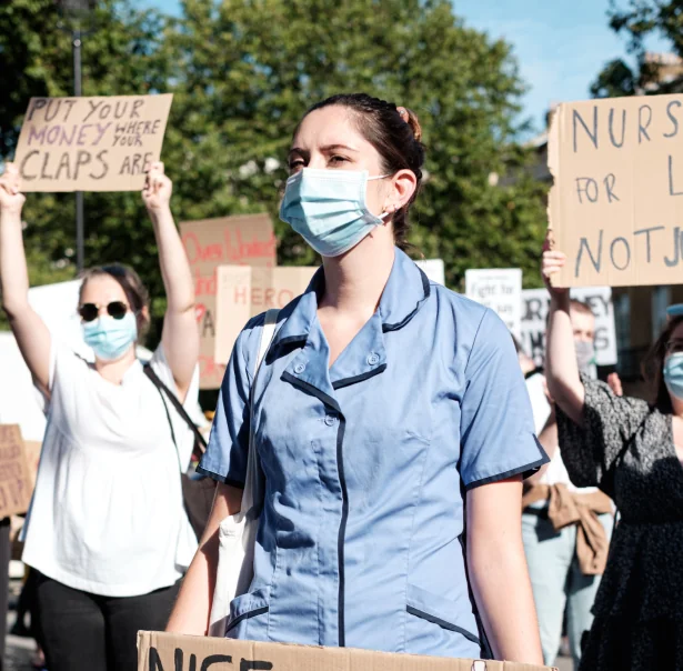 Healthcare workers wearing masks and holding up placards. is backing a petition calling for all NHS healthcare workers to be offered a free Covid booster vaccine this winter.